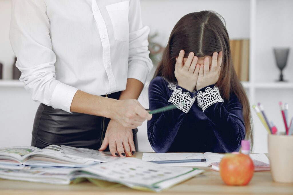 Little schoolgirl covering face with hands and crying while unrecognizable woman persuading to do homework and writing in copybook in modern workplace